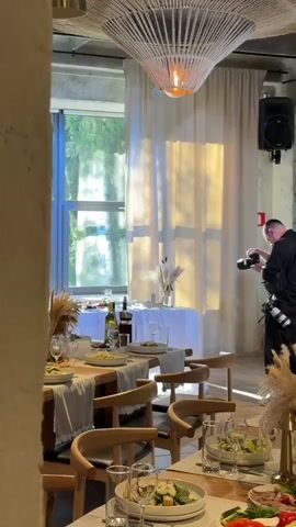 A man wearing a black shirt and black pants is holding a camera and taking pictures. He is standing in a room that appears to be a restaurant or event space. The room is decorated with tables set for a meal, with plates, glasses, and bottles of wine. There are also decorative arrangements of dried grasses on the tables. A large window with white curtains is visible in the background, with sunlight streaming through. A speaker is mounted on the wall above the window. The man is focused on his photography, with his back partially turned to the camera.
