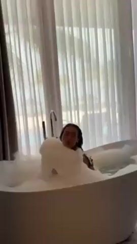 A woman is in a bathtub filled with bubbles. She is wearing a bathing suit. The bathtub is white. Behind her is a large window with white curtains, showing an ocean view with palm trees. She turns her head away from the camera.