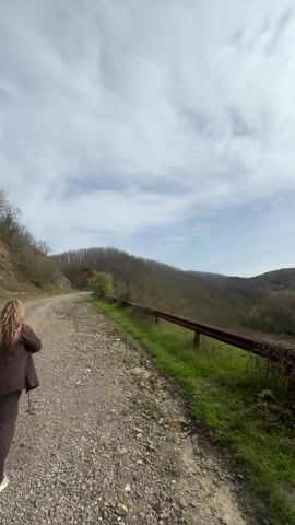 The video shows a gravel road running alongside a rusted metal guardrail. Beyond the guardrail, a view opens up over a green valley with trees and hills in the background. The sky is cloudy, but the sun is shining brightly through the clouds. The video is taken from a ground-level perspective. There is no visible human presence in the shot.