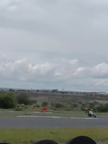 A person riding a small white and yellow motorcycle travels along a paved track on a cloudy day. The landscape around the track is grassy with sporadic trees and a distant cityscape. As the first rider passes, a second person on a green and black motorcycle comes into view. The edge of the track is marked by stacked black tires and there are banners in the background.