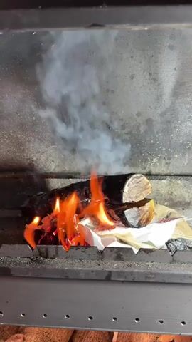 The video shows a close-up, medium shot of a log fire in a fireplace. The scene is dimly lit, suggesting it may be evening or nighttime, or simply the ambient light within the fireplace. The central focus is a split log resting on some crumpled white paper and what appears to be a brown paper bag, which are also burning. Flames in vibrant oranges and yellows lick at the wood and paper, producing wisps of white smoke that rise and dissipate against the dark, rough interior of the fireplace. The log itself has a rough, textured bark and shows the grain of the wood where it has been split. The metal grate of the fireplace is visible in the foreground, with small, evenly spaced holes. Below the grate, more logs are visible, stacked and ready to be burned, hinting at a cozy and warm indoor setting. The overall impression is one of warmth and domesticity, with the crackling fire providing light and heat. There is no visible text in the video. The location context of "Столбище, Russia" suggests this is likely a home fireplace in that region.