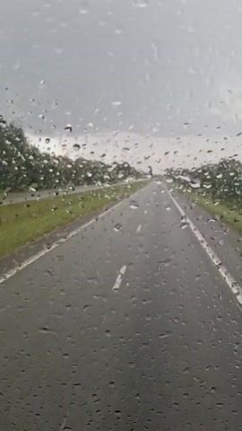 The video shows a view through a windshield covered in raindrops, looking out at a wet, gray highway. The road is dark asphalt with white lane markings. On either side of the road, there are grassy shoulders and trees. The sky is overcast and the overall lighting suggests it is daytime and raining. The perspective is from a moving vehicle, likely a car, looking forward. There are no people visible in the video.