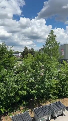 The video shows an outdoor view, possibly from a window or balcony. The sky is blue with fluffy white clouds. In the foreground are several black garbage bins and a small blue car parked nearby. Beyond the bins and car, there are lush green trees and shrubs. In the background, there is a commercial building with a sign that reads "boulderwelt" in a modern font. Below that sign are smaller signs for "REWE", "ALDI", and "dm". The camera pans slightly to the right, focusing on the building in the background. The scene appears to be set in a suburban or semi-urban area, with a mix of nature and commercial development.