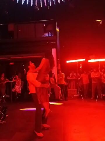 In a nightclub, a man holds a woman in the air, her arms outstretched. He rotates in the center of a dance floor, illuminated by red and blue lights. Several people sit and stand around the dance floor. There's a DJ booth behind the dancers and a bar area to the left with people seated at tables. Some individuals are holding up phones to record.