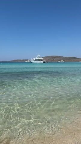 This is a vertical video of a tropical beach with crystal clear turquoise water. A large white yacht is anchored in the distance, with two smaller boats nearby. The water is shallow and clear, revealing the sandy bottom. The sky is a clear blue, with a few faint clouds on the horizon. The overall scene is serene and idyllic.