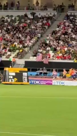 The video shows an athletics stadium with a crowd watching a relay race on the track. The runners are passing the baton. There are advertising boards for Seiko, Honda and other companies.