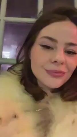 A woman is recording a video of herself in front of a window. The woman has fair skin, brown hair, and pink lipstick. She is wearing a white, furry jacket. Her hair is shoulder-length, and she has winged eyeliner. The video appears to be taken at night. Outside the window, there is a blurry view of streetlights and buildings, lit with purple light. The woman is talking, and her facial expressions vary from a grimace to a smile. In the background, an intermittent rumbling can be heard.