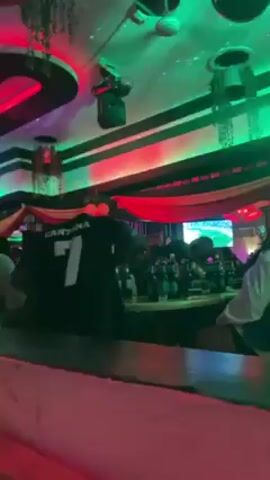 The video shows a crowded bar. A person wearing a black jersey with the name "Cantona" and the number "7" on the back is standing at the bar. A TV screen is visible in the background, displaying a soccer game. Several bottles are lined up on the bar, and patrons are seated around a table. The bar is decorated with colorful lights, including red, blue, and green. There is also some fabric draped across the ceiling.