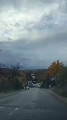 The video shows a car driving down a street in a residential area. The road is paved and wet, and there are trees with autumn foliage on either side. In the background, there are houses and hills, and the sky is cloudy. Another car is driving towards the video recorder.