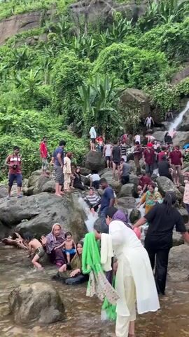The video shows a busy natural scene with a waterfall cascading down a rocky incline. A large group of people are present, many of them dressed in casual clothing, with some wearing traditional attire. They are spread across the rocks and in the water, engaged in various activities. Some individuals are sitting or standing on the rocks, observing the waterfall or interacting with others. Several people are in the water, seemingly enjoying the coolness and the flow of the waterfall, with some appearing to be bathing or playing. Others are in the process of navigating the rocky terrain, with some assisting each other. The environment is lush and green, with dense vegetation covering the surrounding hills. The overall atmosphere appears lively and recreational, with a sense of community gathering.