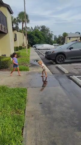 Two young girls are interacting on a wet sidewalk adjacent to a parking lot. The girl on the left, with dark, long hair, wears a purple t-shirt and denim shorts. She appears to be holding a spray bottle. The girl on the right, with lighter, wavy hair, is crouched down. She is wearing a cream-colored t-shirt with colorful hearts and patterned shorts. The ground is wet, creating reflections. In the background, several cars are parked, some covered with car covers. Green foliage and a building are also visible. The overall environment suggests an outdoor, possibly residential or apartment complex setting.