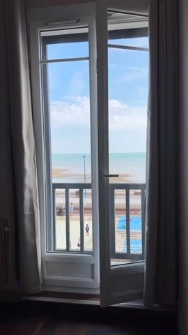 The video shows a view from a room looking out through an open balcony door. Outside, a blue sky with scattered clouds is visible, with the sea beyond. A railing separates the balcony from a promenade or walkway below. People can be seen on the promenade, some wearing casual clothing. There appears to be a swimming pool area with blue tiling to the right of the promenade. The balcony door is white and has glass panels. Light-colored curtains hang on either side of the doorway. The floor inside the room is dark wood. The scene is static, with no discernible movement other than the subtle shifts in light and shadow due to the clouds.