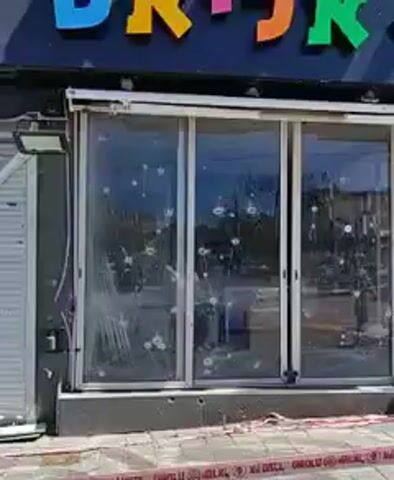 The video shows the exterior of a commercial building that appears to have sustained damage. The building's storefront has glass windows that are shattered with numerous holes, possibly from gunfire. A metal security shutter on one side is significantly damaged, bent and broken. The sidewalk in front of the building is marked with red and white police tape, indicating the area is likely under investigation or restricted. The building has a sign in Hebrew on the top. The surrounding environment appears to be a city street with other buildings and vehicles visible in the background. No people are clearly visible within the immediate frame of the video, although one person is seen in the background. The overall impression is of a scene following a violent incident.