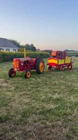 The video pans across a grassy field at sunset. On the left, a red tractor with yellow wheels and accents is parked. To its right, a small, colorful wagon, painted red and yellow with a brown box on top, is hitched to the tractor. Further to the right, a red and yellow combine harvester is connected to another red and yellow wagon. Beyond that, a miniature steam engine, painted black, red, and yellow, is visible. Another red and yellow machine, possibly a smaller piece of farm equipment, is to the right of the steam engine. Finally, on the far right, another tractor, this one white with red and black accents, is parked. In the background, a house, trees, and a telephone pole are visible. The sky is a pale blue, transitioning to orange near the horizon.