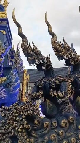 This is a close-up video of a temple or monument with elaborate, dark blue and gold serpent-like statues. Multiple serpentine heads with ornate details and golden crests are visible, along with a gold statue of a person in prayer. The background shows a cloudy sky and parts of a blue building with intricate designs. The camera is static.