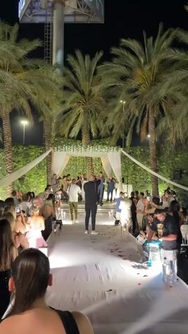 This video captures a nighttime outdoor wedding ceremony at a venue featuring palm trees and lush greenery. A white carpeted aisle is illuminated by spotlights and lined with glass candle holders, some containing lit candles and rose petals. At the end of the aisle, a chuppah, adorned with white drapes, greenery, and fairy lights, serves as the focal point. Several people, presumably the wedding party and officiant, are gathered under the chuppah. The officiant, dressed in a dark suit, appears to be speaking into a microphone. To the left of the chuppah, a band or musicians are visible. The ceremony is being documented by photographers, one of whom, wearing a black shirt and holding a camera, is positioned in the foreground, facing the chuppah. Guests are seated on both sides of the aisle, many holding up phones to record or photograph the event. The overall atmosphere suggests a joyous and celebratory occasion.