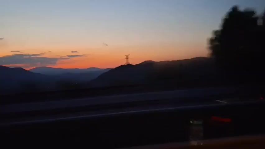 The video shows a blurry view from a moving vehicle at dusk. The sky is a gradient of dark blue at the top, fading into orange and pink hues near the horizon, where mountains are silhouetted. Dark trees are visible on the right side of the frame, also appearing blurred due to motion. A dark road with white lines is in the foreground, suggesting the vehicle is on a highway. The tail lights of a passing truck are briefly visible on the right side of the road, indicated by streaks of orange light. The overall impression is of traveling through a scenic landscape as daylight fades.