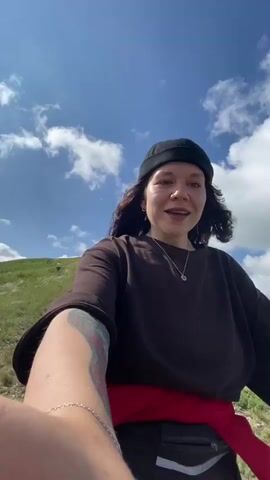 A medium shot shows a young woman with dark curly hair wearing a black beanie and a brown long-sleeved shirt over a red hooded sweatshirt. She is standing on a grassy hillside with a scenic view of rolling green hills and a bright blue sky with white clouds in the background. Her left arm is extended toward the camera, suggesting she is filming herself. She is smiling and looking slightly off to the side. In the background, other people are visible on the hill, walking and gathered together. The overall impression is of a casual outdoor activity or hike.