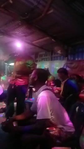 The video appears to be taken inside a small, dimly lit bar or club. There are at least four people visible. One person stands and seems to be singing into a microphone, and the other three are seated at tables. The room has a corrugated metal roof and what appear to be makeshift decorations. Colorful lights flash around the room.