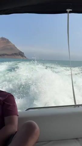 The video shows the wake of a boat moving through turquoise water under a clear blue sky. In the distance, a large, rocky island with a small structure on top is visible. The camera is positioned from inside the boat, looking towards the stern, where a person wearing a maroon t-shirt is seated on the right side of the frame. The boat appears to be a speed boat with white upholstery. The scene suggests a sunny day, likely daytime, given the brightness and clear sky. The environment is coastal, with the island hinting at a location near land. The churning water indicates the boat is moving at a good speed. No visible text is present in the media. The location context provided is Callao, Peru, which aligns with a coastal setting and the appearance of the island. The overall mood is one of recreation and enjoyment of a boat trip.