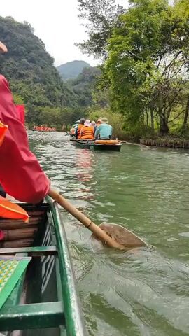 A person in a red jacket is rowing a small boat on a river. The water is green and there are other boats filled with people wearing life jackets in the distance. The person is using a bamboo paddle with a white plastic blade. The river is surrounded by trees and hills. The sky is overcast.