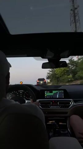 The video shows the interior of a car from a passenger's perspective, looking forward. A person wearing a white baseball cap is driving, with their hands on the steering wheel. The car's dashboard displays a digital speedometer and a navigation screen showing a route. Outside the car, the landscape appears to be rural, with trees and a road. Another car is visible ahead on the road. The sky is overcast, suggesting it is either early morning or late evening.