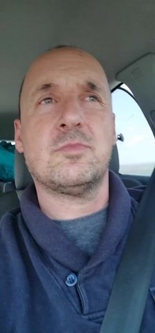 A man in a car is taking a video of himself while driving. He is wearing a navy-blue sweater over a gray shirt and a seatbelt. He has short, thinning hair and a trimmed beard. A young girl sits in the back seat, looking ahead. There are items piled behind her seat, including a turquoise jacket, pink jacket, and gray and purple car seat. The music is playing loudly. The man glances and smiles at the girl in the back seat.
