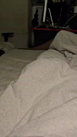 The video shows a person lying in bed under a white patterned duvet. A desk with a computer and other electronic devices is visible in the background. The room appears dimly lit. The camera is stationary.