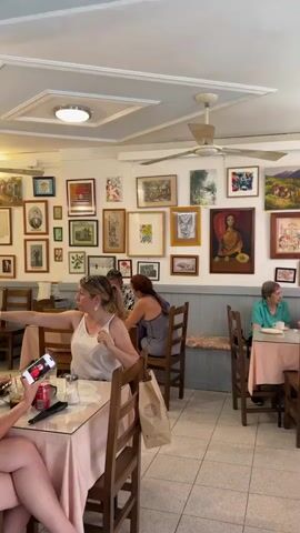The video shows the interior of a restaurant with several patrons. The walls are covered with framed artwork, and there is a ceiling fan visible. In the foreground, a woman wearing a white tank top is seated at a table with a pink tablecloth, holding a smartphone. Another person's legs are visible at the same table. To her right, a woman with dark hair wearing a gray tank top is seated at another table. Further back, a woman in a green top is seated at a table. To the left, a display case with bottles of drinks is visible. The overall impression is of a casual dining establishment.