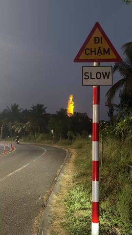 The video shows a curved road at dusk with a large, lit statue of a person visible through the trees in the distance. A signpost stands beside the road, displaying a red triangle with the words “DI CHAM” on it, and a white rectangle with the word “SLOW” underneath. The signpost is striped red and white. The road curves to the left, and the grass beside it is green. The sky is dark, and the statue is illuminated in a yellow-orange light.