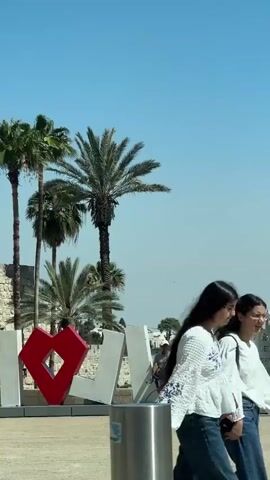 A vertical, low-angle, medium shot captures a bright, sunny day with a clear blue sky above. In the foreground, a large, white, three-dimensional sign reads "I LOVE JERUSALEM" with a bright red heart in place of the word "love." The letters are large and appear to be made of metal or a similar sturdy material, with the "I" and the "heart" on the left, followed by "L," "O," "V," and "E." The letters are slightly tilted forward.

A man wearing a white t-shirt, blue jeans, and a white baseball cap is seated on the "V" of the sign, leaning back with his legs crossed. He appears relaxed and is looking downwards. To the right of the sign, another person, likely a male, is walking away from the camera, carrying a red bag on his right shoulder.

In the background, several palm trees are visible, with their fronds reaching towards the sky. Beyond the trees, the cityscape of Jerusalem unfolds, featuring several buildings with domed roofs and towers, including what appears to be a church or religious structure. The overall scene is one of a tourist landmark, bathed in natural light.