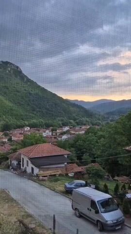The video captures a peaceful village nestled in a lush green valley. Rolling hills covered in dense forest form a dramatic backdrop to the cluster of houses with orange-tiled roofs. A few vehicles, including a silver van and a car, are parked on a paved road in the foreground, suggesting a quiet, lived-in atmosphere. The sky above is a canvas of soft clouds tinged with the warm hues of either sunrise or sunset, casting a gentle glow over the entire landscape. The overall impression is one of tranquility and natural beauty.