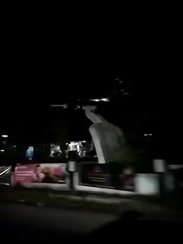 The video shows a view from a moving vehicle at night. The scene is dimly lit, with some buildings and streetlights visible. There is a large, gray structure in the background that appears to be under construction or renovation. In the foreground, there is a billboard with pink and white coloring, featuring images of people. The vehicle appears to be driving on a paved road. The overall impression is of a semi-urban or rural area at night.