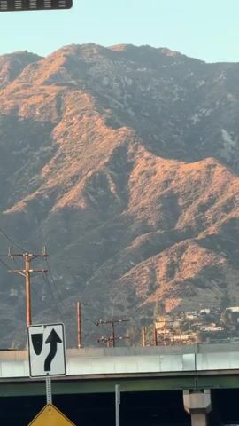 The video pans across a mountainous landscape illuminated by warm sunlight. In the foreground, there is a highway overpass with traffic signs, including one indicating a clearance height of 15 feet 3 inches. Telephone poles and wires are visible along the side of the road. Houses are scattered on the lower slopes of the mountains. The sky is clear and blue.