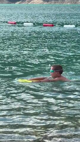 A young boy wearing goggles and swimming in a lake holds a yellow kickboard. He is positioned in the middle of the frame, with his body angled slightly to the right. The water is a turquoise green, with ripples and small waves visible. In the background, a line of buoys, alternating red and white, stretches across the water. Further in the background, the shoreline is visible, appearing as a rocky, tree-covered slope. The lighting suggests it is daytime.