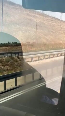 This video depicts a view from inside a vehicle, likely a bus or van, looking out through the windshield. The scene is a highway passing through a dry, hilly landscape under a bright, sunny sky, suggesting a warm, possibly hot, day. The terrain is arid, with sparse vegetation and a large, barren hillside dominating the background. Power lines and a lamppost are visible on the hillside, indicating infrastructure in the area.

The highway itself has guardrails separating it from the adjacent land. On the left side, beyond the guardrail, is a field of dry, stubbly vegetation. The road appears to be well-maintained. The overall impression is of a journey through a somewhat desolate but naturally scenic environment.

A prominent detail in the video is a blue road sign with white text, which is visible on the right side of the highway. The text on the sign appears to be "a.ea", though it is partially obscured and distorted by reflections on the glass. This sign might indicate a town, exit, or geographical feature.

The video captures a sense of movement as the vehicle travels along the road. The reflections on the windshield suggest the presence of the vehicle's interior, including possibly seats and parts of the dashboard. No people are explicitly visible, but the perspective implies someone is operating the camera from within the vehicle. The mood is calm and observational, fitting for a travelogue or a snapshot of a journey. The location context provided, Bnei Shimon Regional Council, Israel, aligns with the dry, hilly terrain depicted.