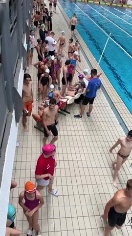 The scene depicts a large indoor swimming pool area filled with people. A group of young swimmers, mostly wearing swimsuits and caps, are gathered near the side of the pool, talking and waiting. Some are standing, while others are sitting on reclining chairs. A man in a blue shirt appears to be addressing them. A few people are walking along the tiled deck surrounding the pool, and others are already in the water, swimming in the lanes. The pool itself is a bright blue, divided into marked lanes. The area is well-lit and appears to be a competitive swimming facility. The video seems to be taken from an elevated viewpoint.