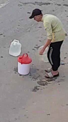A person wearing a light-colored shirt, dark pants, and a black baseball cap is holding a red and white container with a handle. There are two large white containers with handles on the ground near the person. The person is walking on a gray, uneven surface, possibly a road or pathway, with some debris and wet patches. Another person in pink clothing walks past in the background.