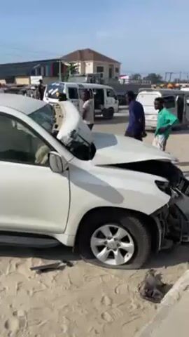 The video shows a white Toyota Land Cruiser Prado that has been severely damaged in a car accident. The front end of the vehicle is smashed in, with the hood buckled and the bumper detached. The vehicle is parked on a sandy area. A flat tire is on the front left. A gray-blue, cab-over, cargo truck with severe front end damage is parked a short distance away. Several people are standing around the car, including a person in a green top and shorts. The accident appears to have occurred on a busy street, as there are other cars and trucks visible in the background. The sky is blue with a few clouds.