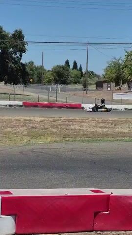 The video shows a go-kart race. The go-kart is white and red with the number 454 on the side. The driver is wearing a helmet and a black and red racing suit. The race is taking place on an outdoor track with a fence and trees in the background. The track is made of asphalt and has red and white barriers. The sky is blue and the sun is shining. The go-kart is moving at a high speed.