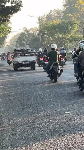A vertical video shows a moderately busy road with several motorbikes and a truck. The sun appears to be rising or setting, casting a warm glow on the scene. Trees line the left side of the road, and more foliage is visible in the background. The road surface is asphalt. On the left side of the road, a white truck is traveling, carrying what appears to be stacked materials on its flatbed. Numerous motorbikes are also visible, all traveling in the same direction. Most of the riders are wearing helmets, and some have passengers. In the foreground, two motorbikes are prominently featured, one dark colored and one light colored. A green bus is seen on the right side of the road, traveling in the same direction as the motorbikes. The overall impression is of daily traffic in a moderately developed area with significant vegetation.