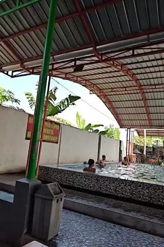 The video shows an outdoor swimming pool area with a covered roof. In the pool, several people, appearing to be children or young adults, are swimming and playing. A few people are also sitting on the edge of the pool. A trash can is visible near the edge of the pool. There are also some plants and a wall in the background. The overall scene depicts a recreational setting, likely a public pool or resort.