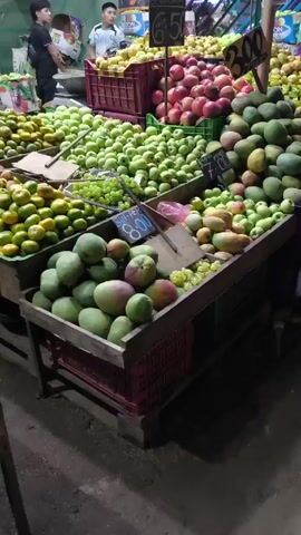The video shows a well-stocked fruit stand in a market.  The camera slowly pans right across multiple crates and displays filled with various fruits: green apples, mangoes (some green, some with red blush), red apples, yellow fruits that may be plums or loquats, grapes (both green and yellow varieties).  Prices are displayed for some of the fruits on small signs (e.g., 8.00, 3.00, 7.00, 1.00, 6.50, 2.00), presumably in a local currency.  Two people are visible in the background on the left of the frame, one wearing a dark t-shirt and the other a light blue shirt.  Boxes of goods, including what appear to be diapers, are visible in the background.  The floor is concrete and somewhat dirty.