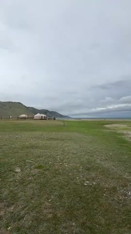 The video shows a landscape view with rolling green hills in the background and a vast grassy plain in the foreground. Several yurts are visible on the plain. The sky is cloudy, with a mix of gray and white clouds. The camera pans across the scene, revealing a body of water in the distance. The water appears calm and reflects the cloudy sky. The overall impression is one of a serene and remote natural setting.