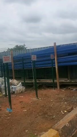 The video shows an outdoor area with a fence and several signs indicating waste segregation. On the left, there are bags of white material, possibly waste, piled up behind a wire mesh fence. A green post with a sign reading "PLASTIC WASTE" is visible. Further to the right, there are more green posts with signs, one appearing to say "FOOD WASTE". In the background, there is a long structure with blue sheeting and some wooden supports. The ground is dirt and gravel. The sky is overcast. The video pans from left to right, revealing more of the area. Towards the right side, there's a larger enclosure with a gate and more debris.