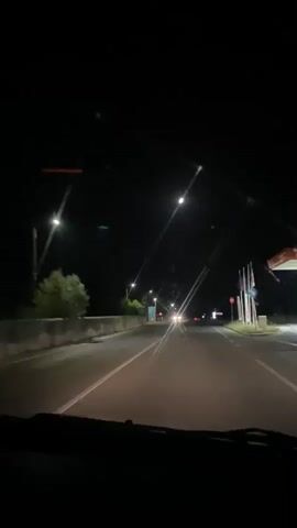 A first-person perspective video shows a person driving a car at night on a paved road with streetlights and other vehicles visible. The camera is mounted inside the car, showing the dashboard and windshield. The road is well-lit by streetlights, and there are multiple lanes. In the distance, the headlights of other cars can be seen. The surrounding environment is dark, with some trees visible on the left side of the road. There are also traffic lights and signs present. The video appears to be a continuous recording without any significant events or actions taking place.