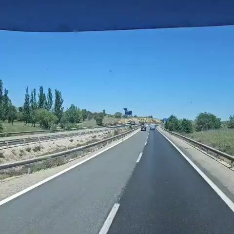 The video captures a view from the inside of a vehicle, presumably a car or bus, traveling on a multi-lane highway on a clear, sunny day. The highway is bordered by metal guardrails, with a dry, grassy landscape adjacent to the road. The most prominent feature in the scene is a large, dark, silhouetted bull statue perched on a small hill overlooking the highway. Several vehicles, including cars and two trucks, are visible on the road. The sky is a clear blue, and there are some trees sparsely scattered across the landscape. The focus of the video seems to be the bull statue, as it is centered in the frame and remains visible throughout the duration of the shot.
