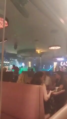 The video shows a dimly lit indoor space, likely a club or bar, with a stage or performance area in the background. Laser lights, primarily blue and green, are projected across the room, illuminating smoke or haze. Silhouettes of people are visible on the stage, and a few patrons are seated in booths in the foreground, some looking towards the stage. One person in the foreground is looking at their phone.