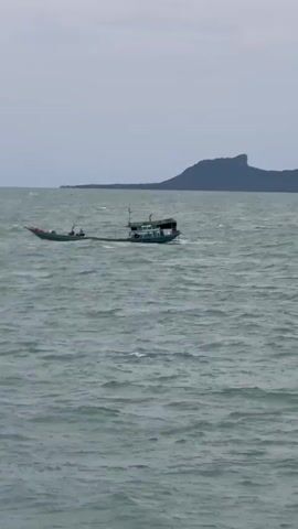 A long, narrow fishing boat with a green hull and a white cabin is moving through choppy, gray water. The boat has several people on board, some of whom are visible near the center of the vessel. In the distance, a dark, mountainous island with a distinct peak rises from the horizon under a hazy, overcast sky. The water's surface is disturbed by small waves, creating a textured effect.