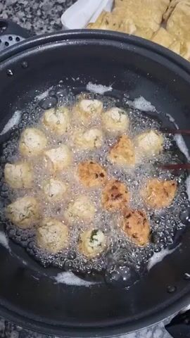 In a close-up, overhead shot, food is being deep-fried in a black non-stick pan. The pan is filled with bubbling hot oil, and numerous small, golden-brown pieces of food are immersed in it. Some pieces are lighter in color, indicating they are still cooking, while others are a deeper brown, suggesting they are nearing completion. Chopsticks are used to stir and turn the pieces in the oil, ensuring even cooking. The food appears to be some kind of fritter or nugget, possibly made of tofu or a vegetable mixture, with visible flecks of green inside some of the lighter pieces. In the background, a white paper towel is visible, and beyond that, a stack of pre-cut, uncooked pieces of what looks like fried tofu or bread are arranged on a light-colored surface. The scene is well-lit, suggesting it could be daytime or under bright kitchen lights. The overall impression is one of a home kitchen setting, likely during meal preparation, possibly for lunch or dinner. There is no visible text in the video.